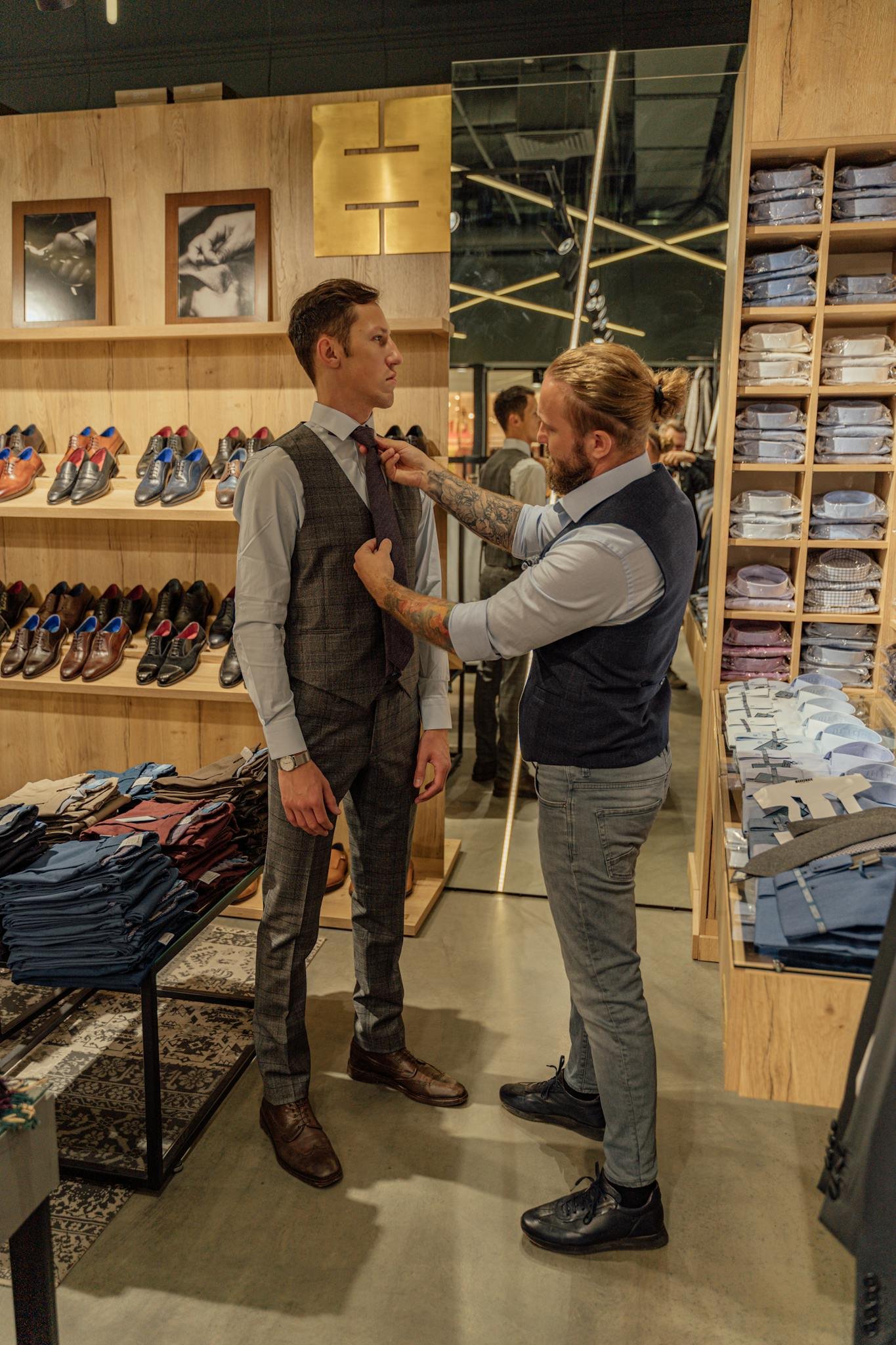 A tailor adjusting a client's tie in a stylish men's fashion boutique with shelves of shirts and shoes around.